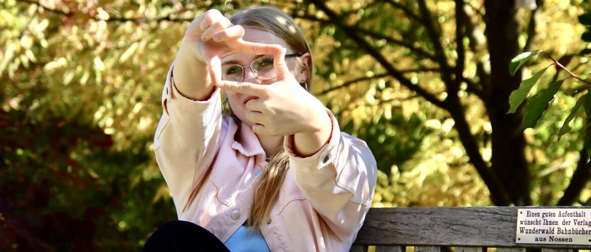 Junge Frau auf einer Holzbank formt mit den Händen einen Bilderrahmen vor dem Gesicht, dahinter goldgelb gefärbtes Laub.