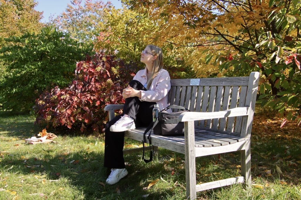 Frau sitzt auf einer Holzbank im sonnigen Garten, umgeben von roten, gelben und grünen Sträuchern und Bäumen, Herbstlaub im Gras.